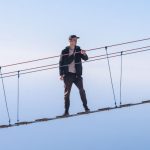 Risk Management - A man skillfully balances on a high rope bridge against a clear blue sky.