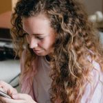 Attic Space - Satisfied female in pink shirt and jeans sitting on bed near suitcases and carton box and browsing cellphone while moving in new apartment