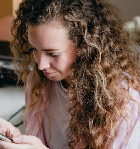 Attic Space - Satisfied female in pink shirt and jeans sitting on bed near suitcases and carton box and browsing cellphone while moving in new apartment