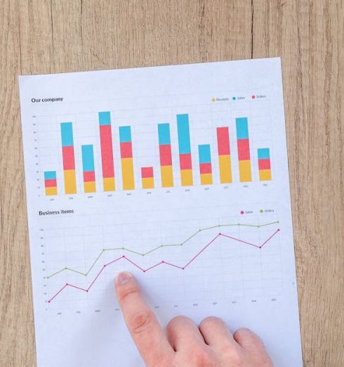 Investors - A hand points to colorful business charts and graphs on a paper sheet on a wooden desk.