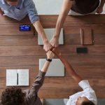 Business Model - A diverse group of professionals engaging in a teamwork celebration with a fist bump over a wooden table indoors.