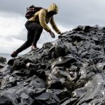 Challenges - Person in yellow jacket climbs black rocky terrain by the ocean under cloudy skies.