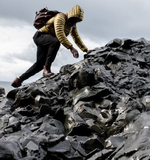 Challenges - Person in yellow jacket climbs black rocky terrain by the ocean under cloudy skies.