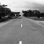 Uncertainty - Black and white photo of a long, straight road lined with trees under a cloudy sky.