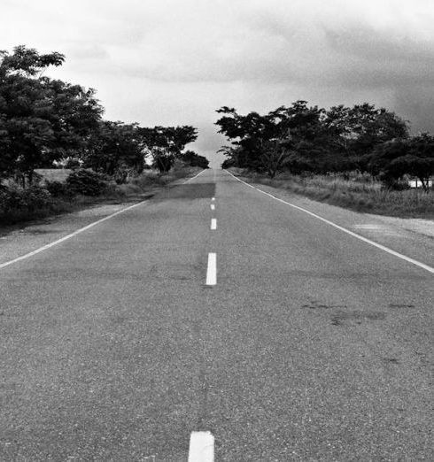 Uncertainty - Black and white photo of a long, straight road lined with trees under a cloudy sky.