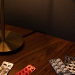 Compliance - Various medication blister packs, pills, and a lamp on a wooden table, creating a moody indoor scene.