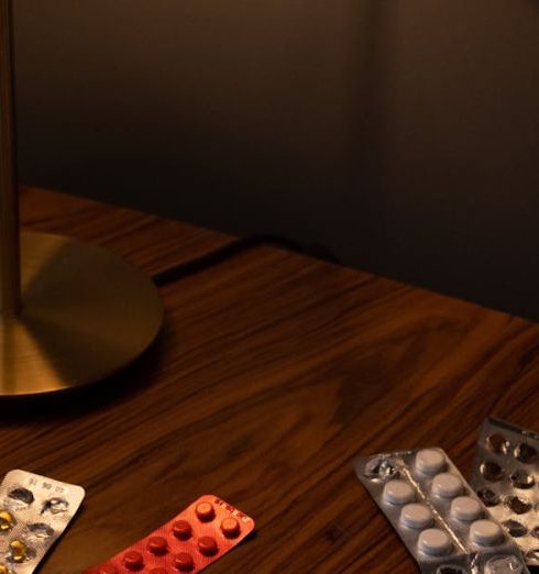 Compliance - Various medication blister packs, pills, and a lamp on a wooden table, creating a moody indoor scene.