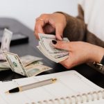 Budgeting - Close-up of person counting cash with notepad on desk, indicating financial tasks.