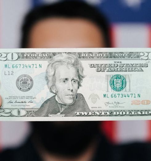 Debt - Close-up of a man holding a 20-dollar bill with an American flag blurred in the background, symbolizing finance and patriotism.