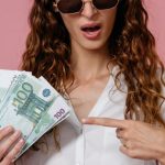 Cash Flow Statements - Young woman in sunglasses holding euro banknotes and pointing, pink background