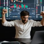 Financial Advisor - Businessman celebrates stock market success with hands raised in excitement at a trading desk.