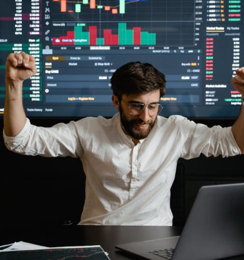 Financial Advisor - Businessman celebrates stock market success with hands raised in excitement at a trading desk.