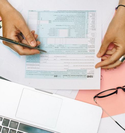 Audit - Hands writing on tax documents with laptop, glasses, and currency on desk.