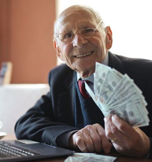 Income - Happy senior businessman showing cash at his desk with a laptop, indoors.