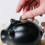 Savings Strategy - Hand inserting coin into black piggy bank among scattered coins, symbolizing savings and financial planning.