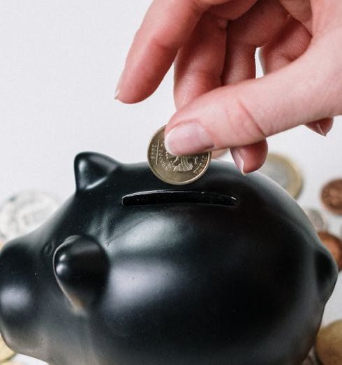 Savings Strategy - Hand inserting coin into black piggy bank among scattered coins, symbolizing savings and financial planning.