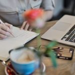 Content Marketing - Woman writing in a notebook with a laptop and coffee cup on a desk. Ideal for workspace inspiration.