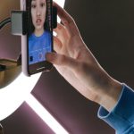 Engaging Content - Asian woman using a smartphone and ring light for video streaming indoors, showcasing modern technology.