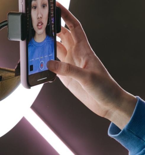 Engaging Content - Asian woman using a smartphone and ring light for video streaming indoors, showcasing modern technology.