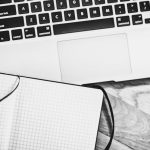 Landing Page - Black and white image of a workspace featuring a laptop, notebook, pen, and coffee cup.