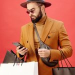 Loyalty Program - Fashionable man with shopping bags using smartphone. Stylish look with beard, hat, and sunglasses.