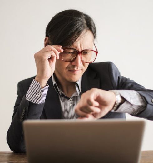 Time Management - Young frowning man in suit and glasses looking at wristwatch while waiting for appointment sitting at desk with laptop