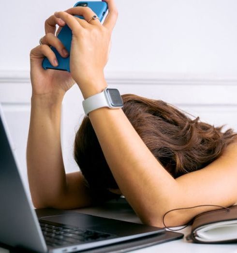 Burnout - A woman overwhelmed by work, resting head on table with laptop, phone, and smartwatch.