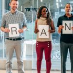 Brand Community - Diverse business team smiling and holding signs spelling 'BRAND' in a modern office.