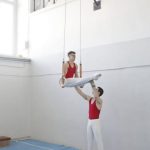 Training - Two male gymnasts engage in a ring routine showcasing strength and balance indoors.