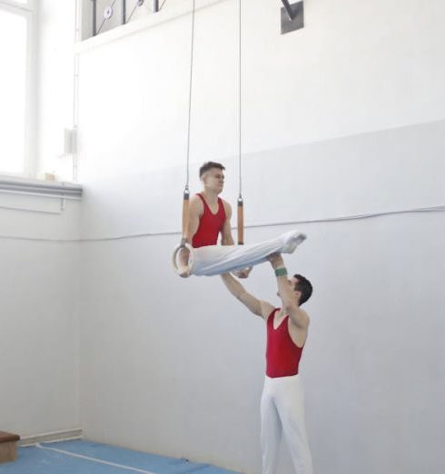 Training - Two male gymnasts engage in a ring routine showcasing strength and balance indoors.