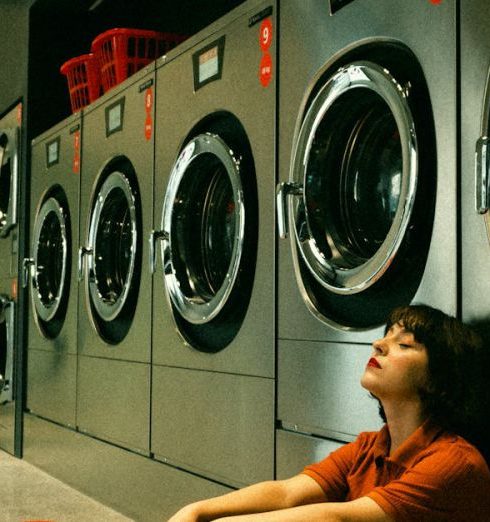 Self-Service - A young woman sitting with eyes closed beside washing machines in a laundromat.